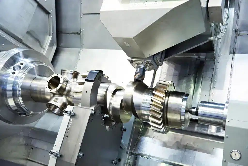 eine CNC Fräsmaschine bei der Arbeit ein Metallstück zu fräsen. Man sieht ein Zahnrad und mehrere Rohranschlüsse