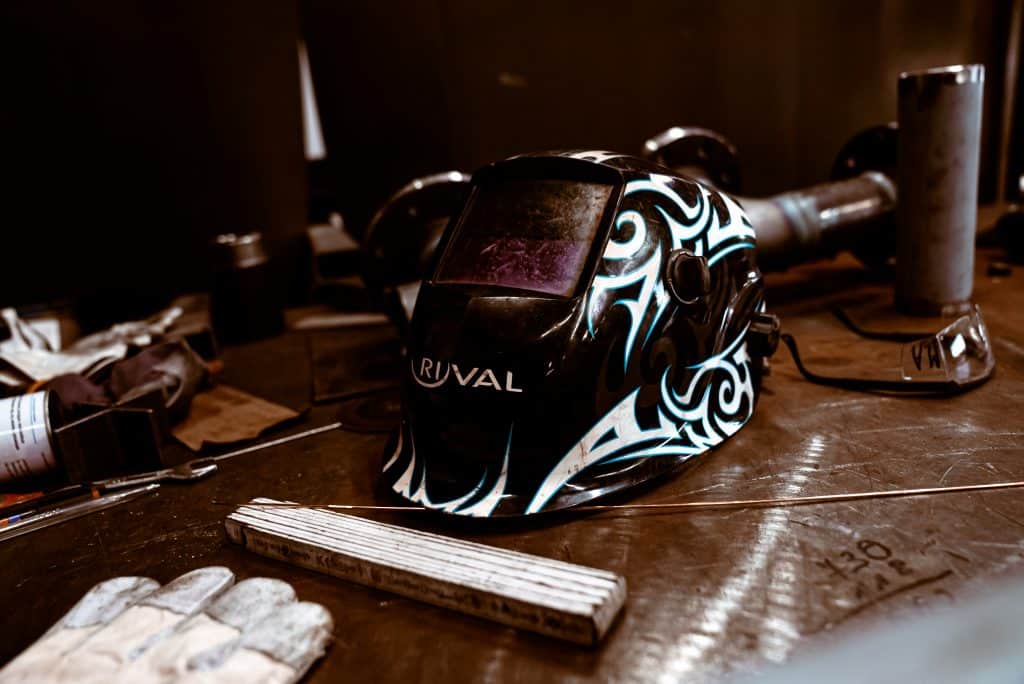 Eine Schweißerschutzmaske mit entsprechender Verglasung liegt auf einem Tisch, zusammen mit Werkzeugen, Schweißerdrähten und anderen Materialien der Industrie