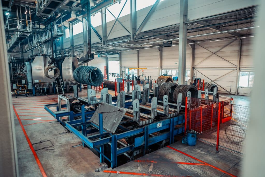 Mehrere Drahtcoils auf mehreren Aufgabetischen in einer Industriehalle, zusätzliche Drahtcoils werden per Lastenkran/Lastaufnahmemittel/Lasthebemittel zu einem Aufgabetisch transportiert