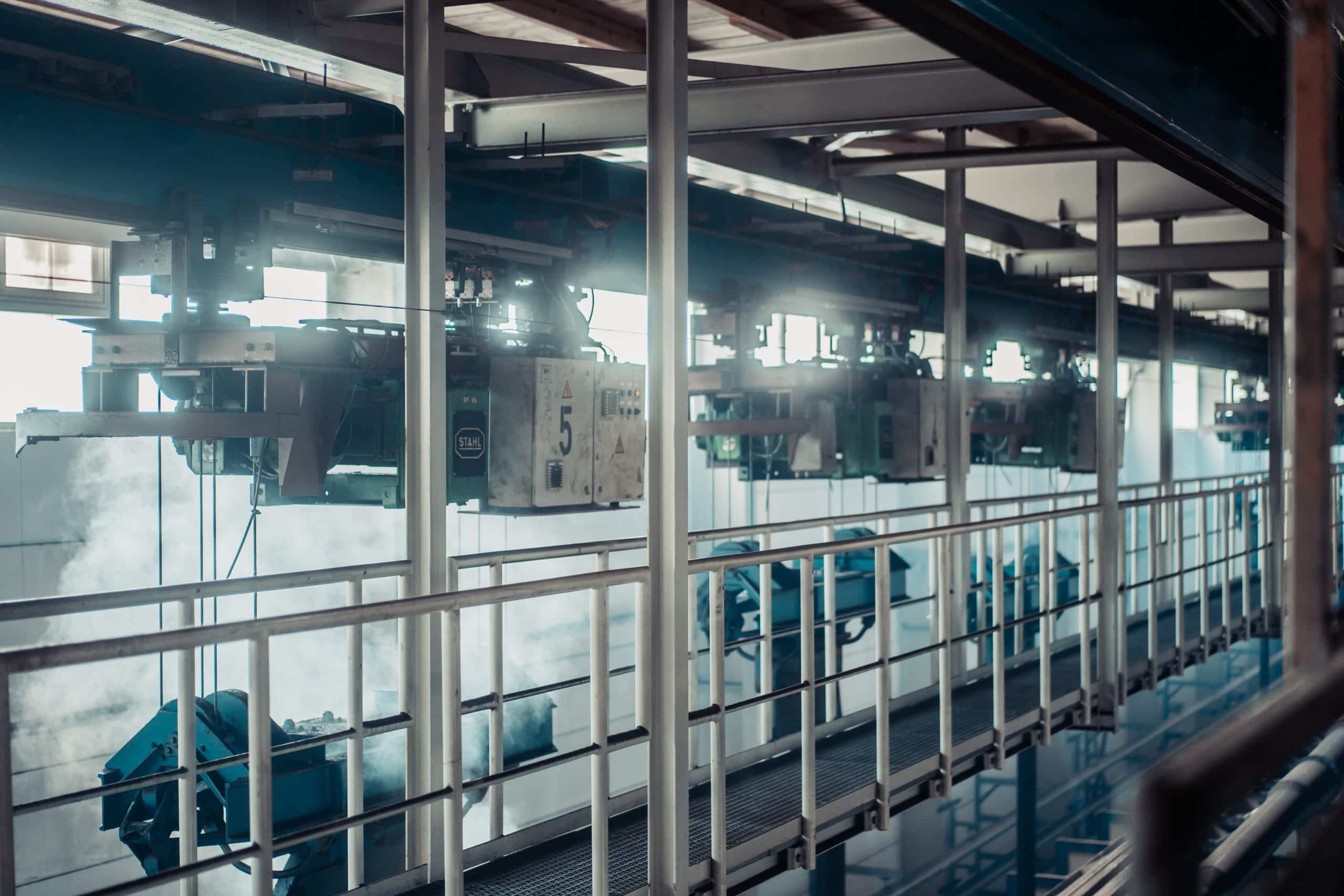 Man sieht einen hoch gelegenen Laufsteg aus Stahl in einer Industriehalle sowie mehrere unter der Decke montierte Kranbahnen. Dies sind gefertigte Lastaufnahmemittel/Lasthebemittel.